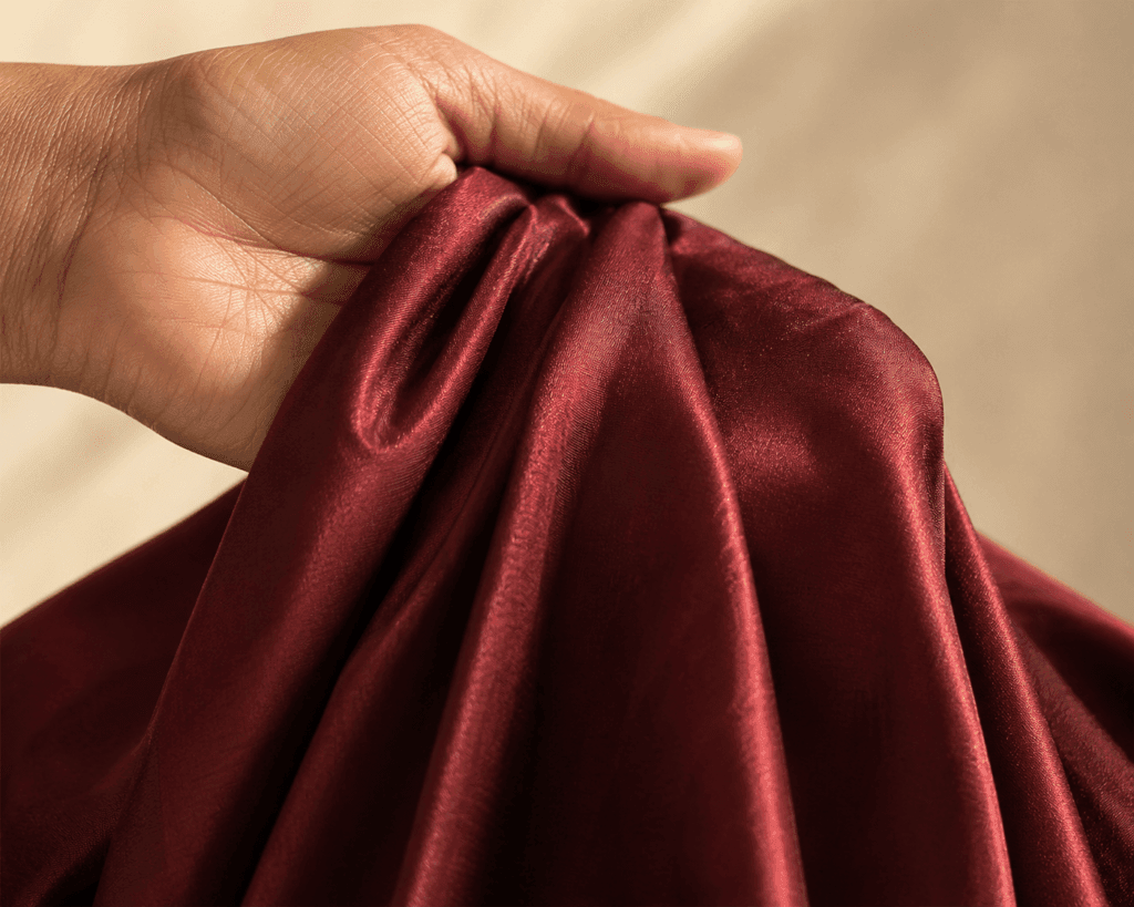 Woman's hand holding pure silk saree fabric to test weight and drape — the silk pools naturally in the palm with characteristic luster and weight.